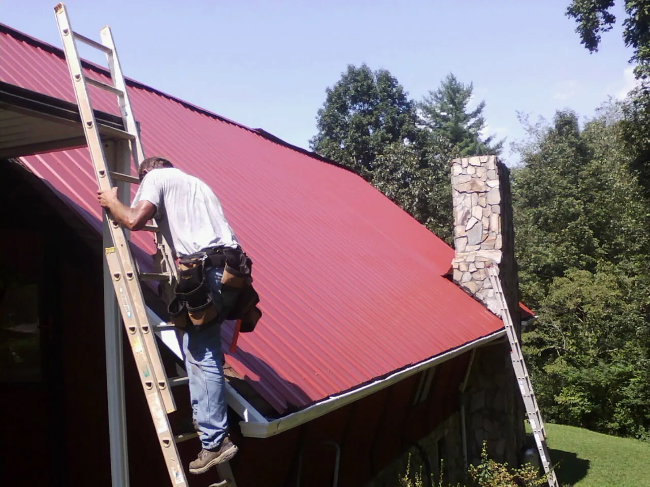 Roofer on a ladder beside a red metal roof and stone chimney — Soffit & Fascia Repair by Roofing Brothers in Carpinteria